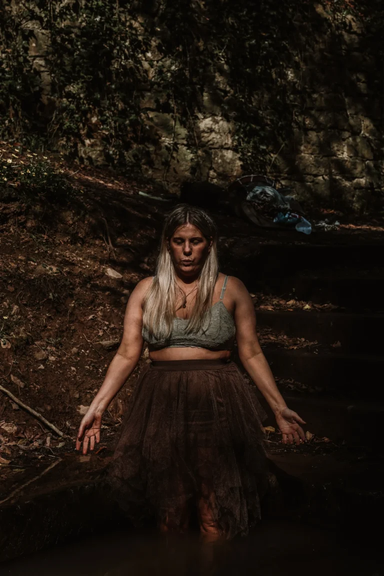 Woman with long hair stands in shallow water, wearing a glittery skirt and green top. Her eyes are closed, conveying a calm, meditative mood.