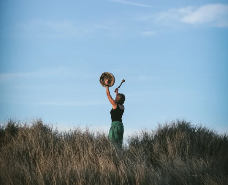A person stands in tall grass holding a drum above their head against a blue sky.