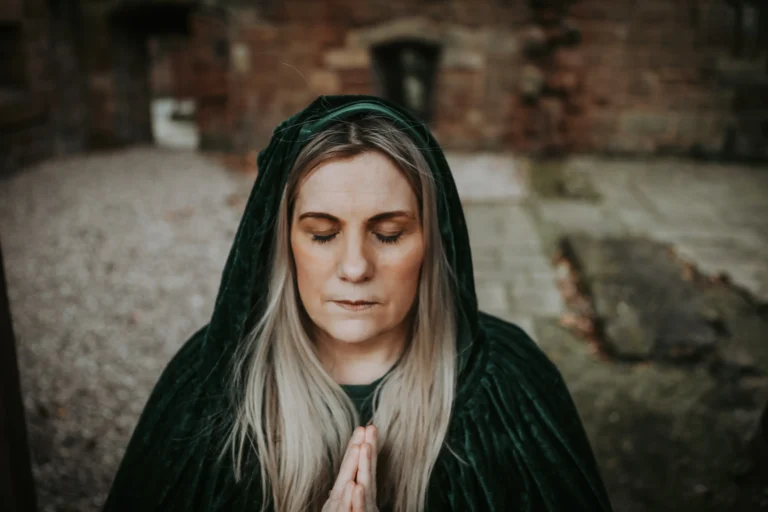 Woman with long blond hair in a green hooded cloak stands outdoors, eyes closed, hands in prayer. The setting includes stone and gravel, conveying tranquility.