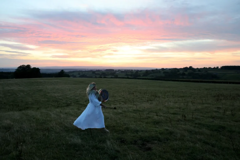 A woman in a flowing white dress walks through a vast field at sunset, playing a drum. The sky is painted with pink and orange hues, evoking tranquility.