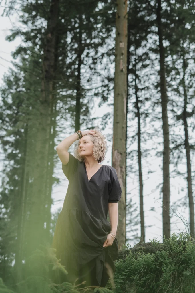A woman in a black dress standing in a forest, looking thoughtfully into the distance.